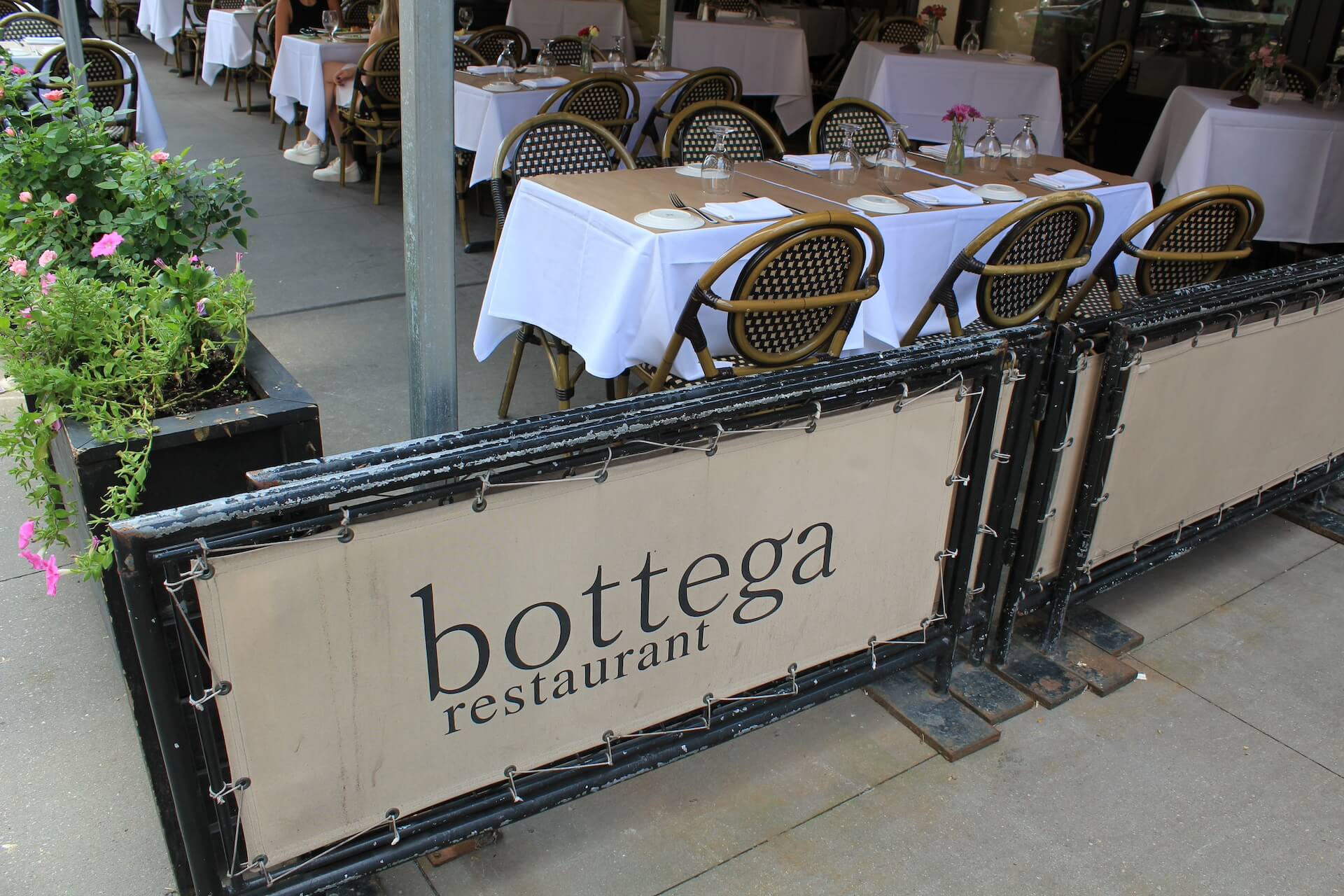 The Bottega Restaurant sign on the sidewalk barrier, with set tables behind it